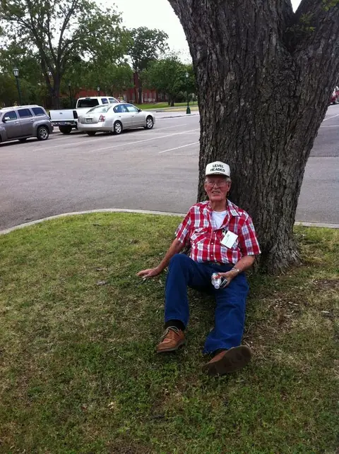 A man in a plaid shirt and cap sitting on the grass against a large tree